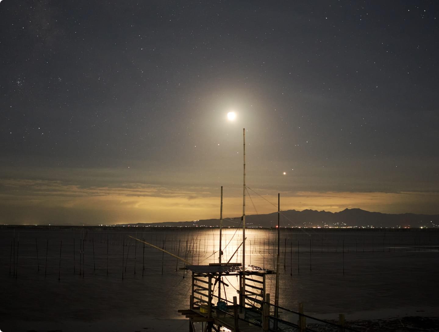 A fishing ground in Okihata River on a full moon night. A traditional four-hook net fishing oar floats on the moonlit water, and the lights of the city spread out in the distance.