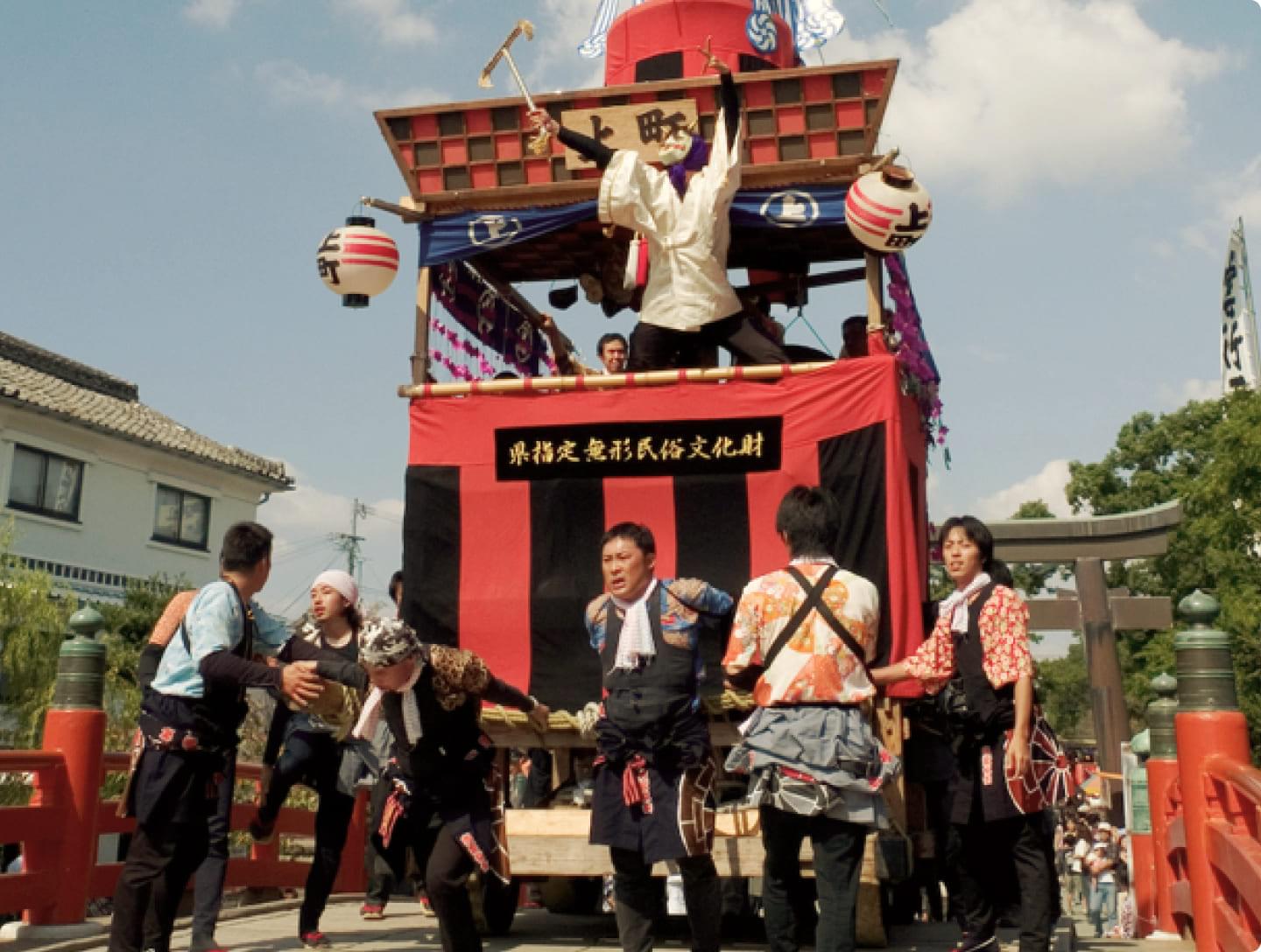 A festival float in Yanagawa. A lively scene of people in happi coats performing traditional routines on a red-decorated float bearing the words "Prefecture-designated Intangible Folk Cultural Property."