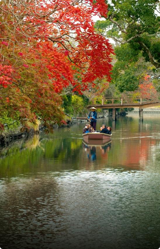 A colorful autumn scene in Yanagawa City. The autumn leaves of the trees are reflected in the water, and red and yellow fallen leaves spread across the riverside promenade. Donko boats quietly move forward, and the cool autumn breeze ripples the water's surface.