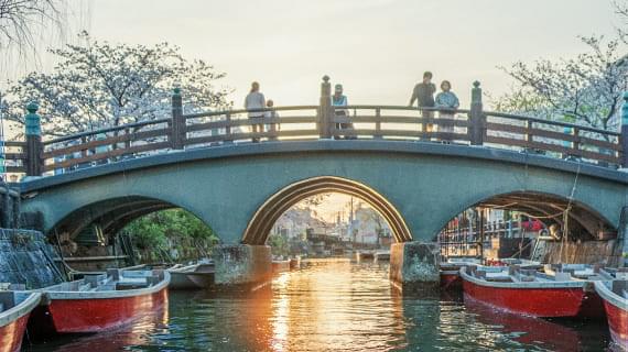 A traditional bridge over Yanagawa’s canals. The sunset reflects on the water, with red sightseeing boats adding to the picturesque scene.