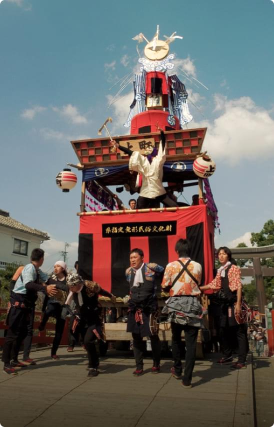 柳川の祭りの山車。県指定無形民俗文化財の文字が記された赤い装飾の山車の上で、法被姿の人々が伝統的な演技を披露している活気ある場面